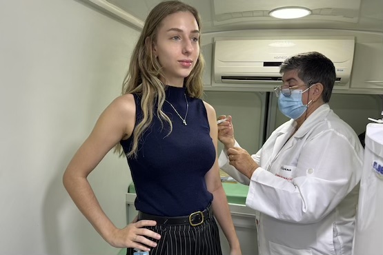 A imagem mostra uma mulher loira, de pele clara, vestindo uma blusa azul escura sem mangas e uma saia listrada preta e branca. Ela está recebendo uma injeção no braço esquerdo de um profissional de saúde. O profissional de saúde está usando um jaleco branco, máscara facial azul e óculos. Eles estão em um ambiente que parece ser uma clínica ou hospital, com paredes claras e iluminação branca.