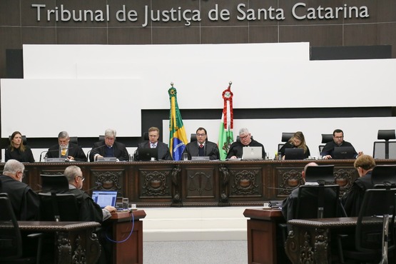 A imagem mostra uma sessão no Tribunal de Justiça de Santa Catarina. Há várias pessoas sentadas em uma bancada elevada, provavelmente juízes ou desembargadores, todos vestidos com trajes formais. No centro da bancada, há duas bandeiras, uma do Brasil e outra de Santa Catarina. A bancada é de madeira escura com detalhes ornamentados. No canto inferior direito, há uma pessoa de costas, também vestida formalmente, olhando para um monitor. No lado esquerdo da imagem, está escrito "Tribunal de Justiça de Santa Catarina".