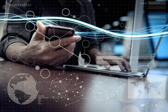 A imagem mostra uma pessoa usando um smartphone com uma mão e digitando em um laptop com a outra mão. A cena está sobreposta com gráficos e elementos digitais, como mapas do mundo, redes de conexões e linhas de dados, sugerindo um ambiente de alta tecnologia e conectividade global. Há também uma linha de luz azul que atravessa a imagem, reforçando a ideia de fluxo de informações.
