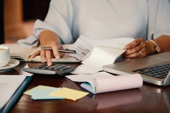 A imagem de uma pessoa usando uma calculadora e fazendo anotações em papéis, com um laptop ao lado. A pessoa está vestindo uma blusa cinza e usa um anel dourado. Na mesa, também há uma xícara de café.