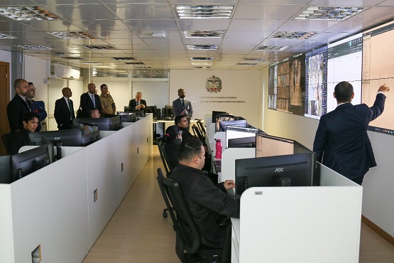 Destaque: A imagem mostra vários homens, alguns sentados e outros em pé, em uma sala de operação, que possui muitas telas. Um deles está em frente a um grande telão, apontando o que está aparecendo e falando para os demais homens que prestam atenção.