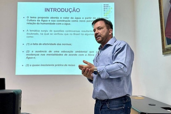 A imagem mostra um homem de pé, aparentemente dando uma apresentação. Ele está vestindo uma camisa social azul e está de perfil, olhando para a direita. Atrás dele, há um projetor exibindo um slide com texto