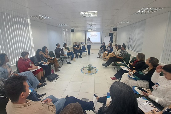 A imagem mostra uma sala de reuniões com várias pessoas sentadas em cadeiras dispostas em um grande círculo. No centro do círculo, há um pequeno arranjo de plantas. Ao fundo, uma mulher está de pé, aparentemente falando para o grupo. Atrás dela, há uma tela de projeção com uma apresentação exibida. A sala é bem iluminada, com luz natural entrando pelas janelas com persianas verticais.