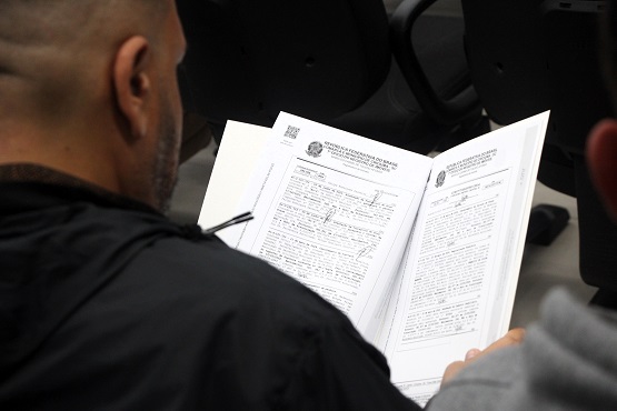 A imagem mostra uma pessoa de costas, com a cabeça raspada, lendo um documento. A pessoa está segurando o documento, que está dentro de uma pasta, com as mãos. Ao fundo, há cadeiras de um auditório ou sala de espera.