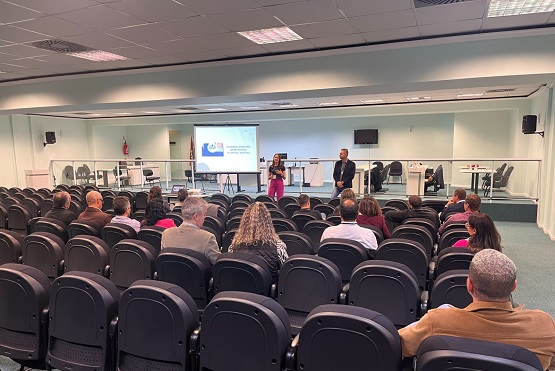 A imagem mostra um auditório em um ângulo amplo, com algumas pessoas sentadas nas poltronas, mais no fundo na imagem o palco com um homem e uma mulher palestrando.