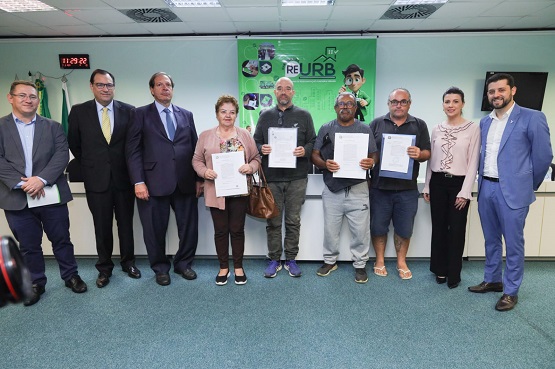 Sete homens e duas mulheres posam para a foto no palco do auditório. Atrás deles há um banner verde chamativo com a escrita "ReUrb. Uma das mulheres e três dos homens seguram um papel com suas duas mãos, o que deve ser um documento.