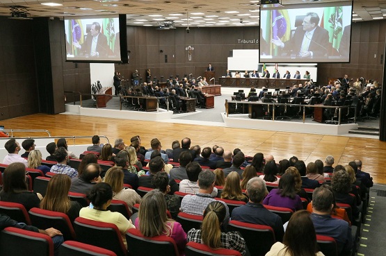A foto é uma imagem ampla do Tribunal, onde aparecem as pessoas que estão assistindo a audiência e a mesa onde estão os juízes e desembargadores. Todas as pessoas se vestem com roupas formais , pendurados no teto há dois telões mostrando imagens do evento.