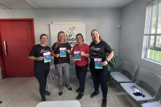 A imagem mostra 4 mulheres segurando um livro colorido. Elas estão dentro de uma sala, atrás delas tem uma porta vermelha, um banner branco com a escrita "Lar Abdon" e uma janela bem iluminada.