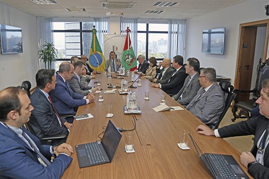 A imagem mostra uma reunião formal em uma sala de conferências. Há uma mesa retangular grande com várias pessoas sentadas ao redor, todas vestidas formalmente. Na cabeceira da mesa, há duas bandeiras: a do Brasil e a de Santa Catarina. A sala tem janelas grandes ao fundo, permitindo a entrada de luz natural. Sobre a mesa, há laptops, copos de água, garrafas de água e alguns papéis.