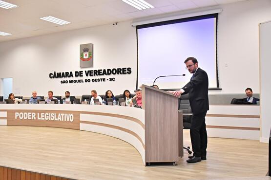 A imagem mostra uma sessão na Câmara de Vereadores de São Miguel do Oeste, SC. No centro da imagem, há um homem de terno preto e óculos falando em um púlpito. Atrás dele, há uma mesa longa com várias pessoas sentadas, possivelmente vereadores. Na parede atrás da mesa, está escrito "CÂMARA DE VEREADORES SÃO MIGUEL DO OESTE - SC" e há um brasão acima do texto. A palavra "PODER LEGISLATIVO" está escrita em uma faixa vertical ao lado esquerdo da imagem.