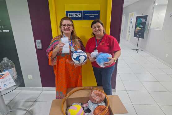 A imagem mostra duas mulheres sorrindo e segurando bolas. A mulher à esquerda está vestindo um vestido longo e colorido, predominantemente laranja, e segura uma bola de vôlei. A mulher à direita está vestindo uma camiseta vermelha e segura uma bola azul e branca. Elas estão em um ambiente interno com piso de azulejo branco. Ao fundo, há uma parede amarela e roxa com uma placa azul que parece ser de uma instituição ou empresa. Há também uma garrafa grande de água sobre uma mesa à esquerda.