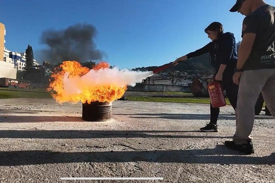 A imagem mostra uma cena de treinamento de combate a incêndio. No centro da imagem, há um tambor metálico com uma grande chama saindo dele, produzindo bastante fumaça preta. À direita, uma pessoa está usando um extintor de incêndio para tentar apagar as chamas. Ela está vestida com roupas escuras e um boné. Ao fundo, é possível ver algumas construções e um céu azul claro. Na parte inferior da imagem, há uma segunda foto menor que mostra outra pessoa segurando um extintor de incêndio, também participando do treinamento.