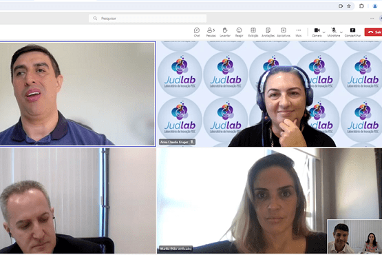 A imagem mostra uma videoconferência com quatro participantes. No canto superior esquerdo, há um homem de pele clara e cabelo curto, usando uma camisa polo azul. No canto superior direito, há uma mulher de pele clara, cabelo escuro preso e usando fones de ouvido. Atrás dela, há um fundo com o logotipo "Judlab". No canto inferior esquerdo, há um homem de pele clara, cabelo curto e grisalho, usando um terno escuro. No canto inferior direito, há uma mulher de pele clara e cabelo castanho claro, solto. No canto inferior direito da tela, há uma pequena janela mostrando uma visão em miniatura de dois dos participantes.