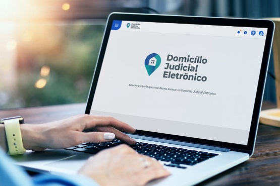 A imagem mostra uma pessoa usando um laptop. A tela do laptop exibe a página inicial de um site chamado "Domicílio Judicial Eletrônico". A pessoa está digitando no teclado com a mão direita. Ela está usando um relógio de pulso amarelo. Ao fundo, há uma janela com uma vista desfocada, sugerindo que a foto foi tirada em um ambiente interno com luz natural entrando.