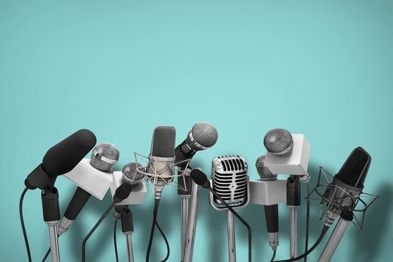 A imagem mostra vários microfones de diferentes tipos e tamanhos, todos montados em suportes. Eles estão dispostos em uma linha, como se estivessem prontos para uma coletiva de imprensa ou uma entrevista. O fundo é de cor azul clara, o que destaca os microfones na frente.