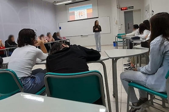 A imagem mostra uma sala de aula com várias pessoas sentadas em carteiras. A foto está orientada na vertical, mas parece que foi tirada de lado, então a sala de aula parece estar deitada.   Na parte superior da imagem, há uma pessoa de camiseta branca e cabelo curto e escuro, sentada de lado, olhando para a frente da sala. Ao fundo, há um projetor exibindo algo na parede.  Na parte inferior da imagem, há outra pessoa de cabelo cacheado e escuro, vestindo uma camisa clara, também sentada em uma carteira.