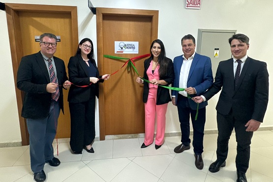 A imagem mostra cinco pessoas em pé, participando de uma cerimônia de inauguração. Eles estão em frente a duas portas de madeira. No centro, há uma fita vermelha e verde sendo cortada, simbolizando a inauguração. As pessoas estão vestidas formalmente, com ternos e roupas sociais. No centro, uma mulher está vestida com um terno rosa. À direita dela, um homem de terno azul segura a fita verde. À esquerda dela, uma mulher de terno preto segura a fita vermelha. Nas portas, há um cartaz com o logotipo da "Justiça Perto". No canto superior direito, há uma placa de "Saída".