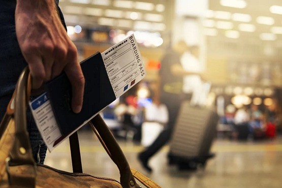 A imagem mostra uma pessoa segurando as alças de uma bolsa de viagem marrom e o passaporte junto com um cartão de embarque em uma mão. A cena parece ser em um aeroporto, pois ao fundo há uma pessoa puxando uma mala de rodinhas e o ambiente tem um aspecto típico de saguão de aeroporto, com iluminação brilhante e movimentação de pessoas.