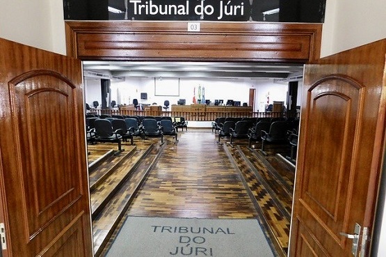 A imagem mostra a entrada do Tribunal do Júri. São duas portas de madeira bem envernizadas e em cima delas há uma placa preta com a escrita "Tribunal do Júri". Na parte de dentro da sala há muitas cadeiras pretas em diferentes níveis, aparecendo os degraus.