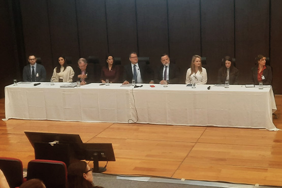 A imagem mostra nove pessoas sentadas atrás de uma longa mesa coberta com uma toalha branca. A mesa está em um palco de madeira. As pessoas estão vestidas formalmente, a maioria em ternos e roupas sociais. Há microfones e garrafas de água sobre a mesa. O fundo é escuro, provavelmente uma parede de madeira ou um painel. Na frente do palco, há algumas cadeiras ocupadas por pessoas assistindo à conferência.