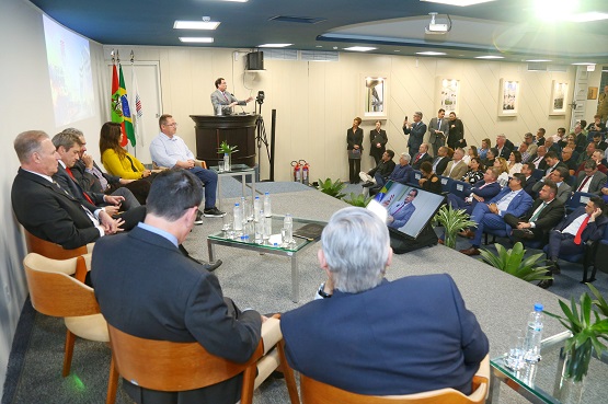 A imagem mostra um auditório com várias pessoas presentes. No centro da imagem, há um palco elevado com uma mesa de centro e várias cadeiras. Sentadas na mesa, há sete pessoas, incluindo homens e mulheres, todos vestidos formalmente. À esquerda, um homem está de pé em um púlpito, aparentemente fazendo uma apresentação ou discurso. No lado direito da imagem, há uma plateia sentada em cadeiras, prestando atenção ao que está sendo apresentado. A sala é bem iluminada e decorada com plantas e bandeiras. Na parede ao fundo, há quadros pendurados.