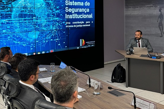 A imagem mostra uma apresentação em um evento. No canto superior esquerdo, há um slide projetado com o título "Sistema de Segurança Institucional" e o subtítulo "Uma contribuição para o sistema de justiça nacional". No canto inferior direito do slide, há um logotipo com as palavras "Justiça do Trabalho".  Abaixo do slide, há um homem sentado em uma mesa, usando um terno cinza e gravata vermelha, falando ao microfone. Ele está olhando para um laptop aberto à sua frente. Na mesa, há também um copo de água e alguns cabos conectados a equipamentos eletrônicos.