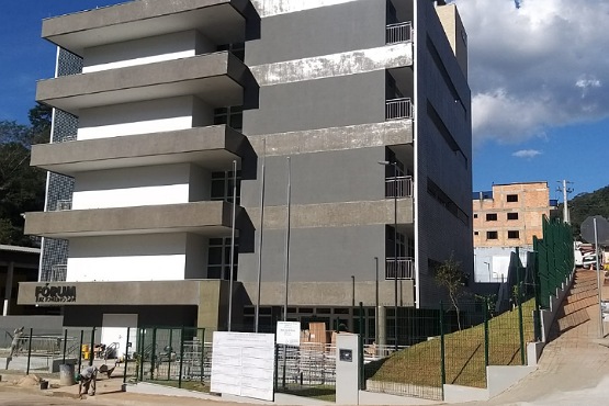 A imagem mostra a fachada de um prédio ainda em construção, em sua frente há alguns baldes e cimento. Ele é marrom e cinza, parece ter três andares e tem algumas sacadas. No fundo da imagem se enxerga o céu, que está azul, e algumas casas. A construção é cercada por uma grade verde.