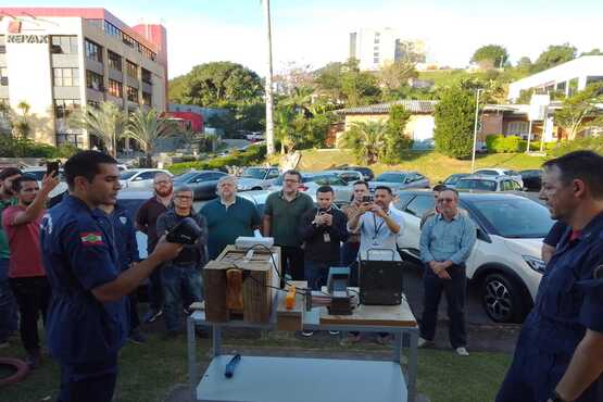 Dois homens vestidos com roupas de bombeiro azul, fazem uma apresentação para um grupo de outros homens que prestam atenção e tiram fotos. Eles estão em um ambiente aberto, atrás deles está a rua, com muitos carros estacionados, há também muitas árvores e um prédio.