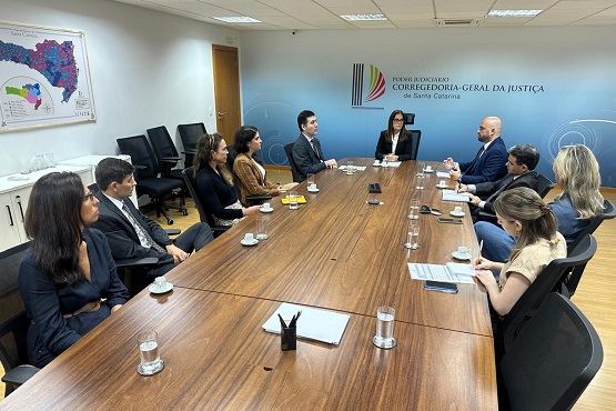 A imagem mostra uma reunião em uma sala de conferências. Há uma mesa retangular de madeira com várias pessoas sentadas ao redor. No lado esquerdo da mesa, há três pessoas, e no lado direito, quatro pessoas. No fundo da sala, há uma parede com um banner que diz "CORREGEDORIA-GERAL DA JUSTIÇA" e um logotipo. Na parede à direita, há um mapa colorido.