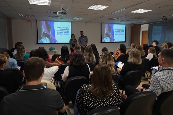 A imagem mostra uma sala com várias pessoas sentadas em cadeiras, assistindo a uma apresentação. Na frente da sala, há duas pessoas em pé, um homem e uma mulher, que parecem estar falando para o público. Atrás deles, há duas telas de projeção exibindo a mesma imagem e texto. O texto nas telas parece ser parte da apresentação, mas não é totalmente legível na imagem. As pessoas na audiência estão prestando atenção, algumas fazendo anotações. A sala é bem iluminada com luzes de teto e tem um ambiente profissional.