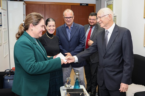 A imagem mostra um grupo de cinco pessoas em um ambiente de escritório. À esquerda, uma mulher de cabelo preso e vestindo um blazer verde está sorrindo e apertando a mão de um homem idoso à direita, que está usando um terno escuro e óculos. Atrás deles, há três pessoas: uma mulher de cabelo preso e vestindo uma blusa preta, um homem de óculos e terno azul, e outro homem de terno escuro e gravata vermelha. No fundo, há uma porta com uma placa de "SAÍDA" e algumas molduras nas paredes. Na frente, sobre uma mesa, há alguns objetos decorativos.
