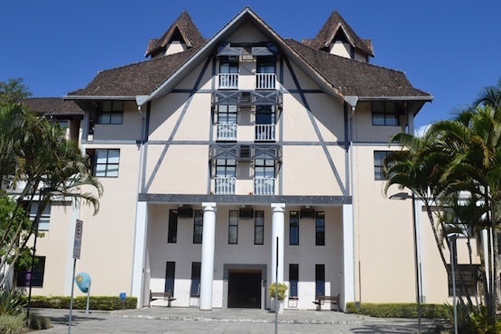 Fórum Central do TJ da comarca de Blumenau. A construção é estilo chaimel com quatro pilares na parte da entrada do local. A imagem também mostra parte do céu com nuvens e algumas palmeiras junto ao gramado verde.