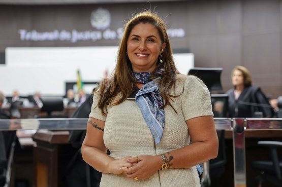 A imagem mostra uma mulher sorridente em um ambiente que parece ser uma sala de tribunal ou uma sala de conferências. Ela está vestindo uma blusa clara e um lenço azul no pescoço. A mulher tem cabelos longos e castanhos. Ao fundo, há outras pessoas desfocadas, possivelmente participando de uma sessão ou reunião. Há também uma tela grande com texto, mas não é possível ler o que está escrito.