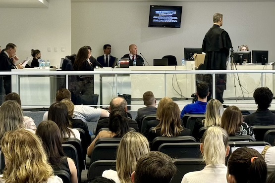 A imagem mostra uma sala de tribunal durante uma sessão. Na parte da frente, há várias pessoas sentadas em cadeiras, assistindo ao que está acontecendo. Na parte de trás, há uma bancada elevada onde estão sentados juízes e advogados. Um homem de terno preto está de pé, aparentemente fazendo uma apresentação ou argumentação. Atrás dele, há uma tela com texto projetado, mas o conteúdo não é legível na imagem. A sala é bem iluminada e parece ser um ambiente formal e organizado.