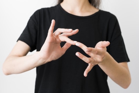 A imagem mostra uma pessoa vestida com uma camiseta preta gesticulando com as mãos, como um intérprete de libras. O fundo da imagem é todo branco.