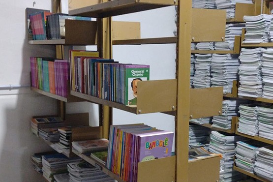 A imagem mostra uma sala com estantes de metal cheias de livros e cadernos. As estantes são de cor bege e estão organizadas em fileiras. À esquerda, há uma estante com várias prateleiras contendo livros de diferentes tamanhos e cores. No centro, há outra estante com livros organizados verticalmente, onde é possível ver as capas de alguns livros, como um chamado "Bambolê". À direita, há uma estante cheia de cadernos empilhados. A sala parece ser um depósito ou uma biblioteca.