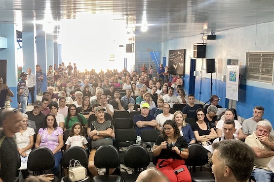 A imagem mostra uma cerimônia em um auditório. Há muitas pessoas sentadas em cadeiras, olhando para a câmera e sorrindo. A maioria das pessoas parece estar prestando atenção ao evento. O ambiente é iluminado e as paredes são de cor azul. No fundo, há um púlpito com um microfone e uma bandeira ao lado. Parece ser um evento formal, possivelmente relacionado à entrega de títulos de propriedade.