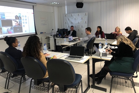 A imagem mostra uma sala de reunião com várias pessoas sentadas ao redor de uma mesa retangular. Há pelo menos oito pessoas visíveis, todas concentradas em laptops ou documentos. Na parede ao fundo, há uma tela de projeção exibindo o que parece ser uma videoconferência com várias janelas de vídeo. A sala é bem iluminada e tem um ambiente profissional, com cadeiras de escritório e mesas brancas. Há também garrafas de água e copos sobre a mesa.