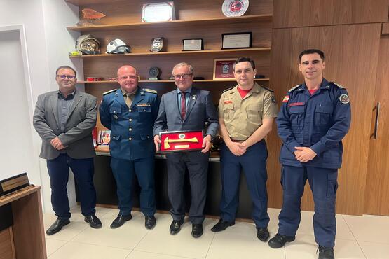 A imagem mostra seis homens posando para uma foto em um ambiente interno. Eles estão em fileira. A maioria deles está usando uniformes militares ou de bombeiros e outros de termos. Ao fundo, há uma estante de madeira com vários objetos decorativos, incluindo capacetes e troféus.