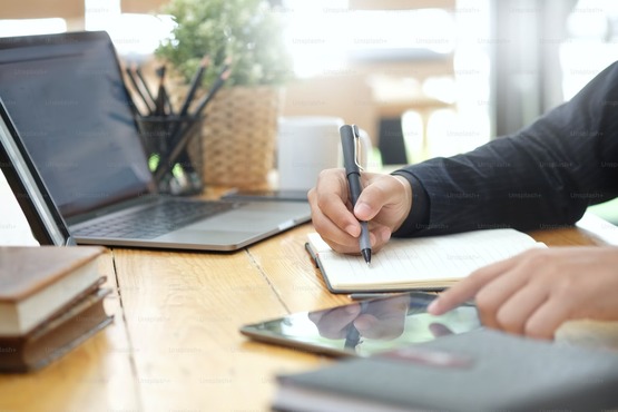 A imagem mostra uma pessoa escrevendo em um caderno com uma caneta. A pessoa está sentada em uma mesa de madeira clara, e ao lado do caderno há um laptop aberto. Ao fundo, é possível ver uma janela com luz natural entrando, e do lado de fora parece haver algumas plantas. A mão que segura a caneta está mais próxima da câmera, enquanto a outra mão está segurando o caderno. A pessoa está vestindo uma roupa escura, possivelmente um casaco ou blazer.