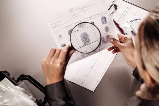 A imagem mostra uma pessoa segurando uma lupa sobre um documento que parece ser uma ficha de impressões digitais. A pessoa está usando um casaco preto e está examinando as impressões digitais no documento com a ajuda da lupa. Há também uma caneta sobre a mesa ao lado do documento. A cena sugere que a pessoa está realizando uma análise ou verificação das impressões digitais.