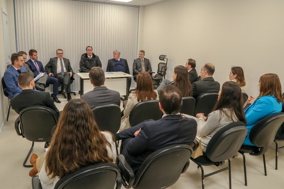 A imagem mostra uma reunião formal em uma sala. Há sete homens sentados em cadeiras ao redor de uma mesa branca. Eles estão vestidos formalmente, usando ternos e gravatas. Atrás deles, há uma parede com persianas verticais fechadas. Na frente deles, há várias pessoas sentadas, de costas para a câmera, também vestidas formalmente. A atmosfera parece ser séria e profissional.