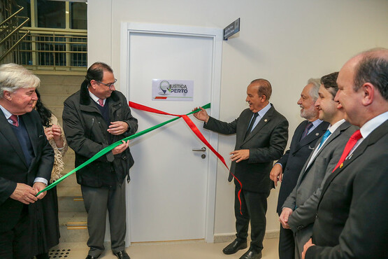 A imagem mostra cinco pessoas participando de uma cerimônia de inauguração. Eles estão em frente a uma porta branca com uma placa que diz "Justiça Perto". Na direita, um homem de terno cinza escuro e gravata vermelha e outro homem de terno cinza claro, estão segurando fitas vermelhas e verdes. Ao lado deles, há uma mulher de cabelos escuros e um homem mais velho de cabelos grisalhos, ambos sorrindo. No canto esquerdo da imagem, há outro homem de perfil, também participando do evento. Todos parecem estar em um ambiente interno.