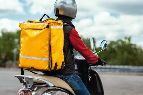 A imagem mostra um entregador de moto visto de perfil. Ele está usando um capacete cinza e uma jaqueta preta com mangas vermelhas. Nas costas, ele carrega uma grande mochila térmica amarela, típica de entregadores de comida. A moto está estacionada, e o entregador está olhando para o lado. Ao fundo, há uma estrada e vegetação desfocada.