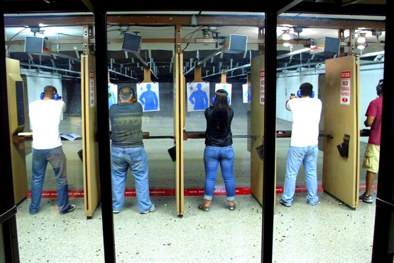 A imagem mostra várias pessoas em um estande de tiro. Elas estão posicionadas em baias individuais, cada uma com uma arma apontada para alvos à frente. Os alvos são de papel e têm a forma de silhuetas humanas. As pessoas estão usando protetores auriculares e óculos de proteção. A imagem está inclinada para a esquerda, o que pode dificultar a visualização.