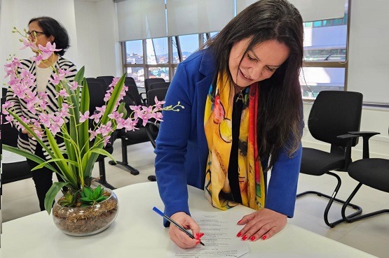 Na foto, uma mulher de cabelos longos e castanhos, vestindo um casaco azul e um cachecol colorido, está assinando um documento sobre uma mesa branca. Ao lado dela, há um vaso de vidro com uma planta de flores roxas. Ao fundo, outra mulher, de cabelos curtos e escuros, está de pé, vestindo uma blusa listrada em preto e branco.