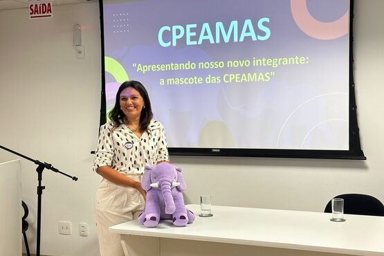 A imagem mostra uma mulher sorridente, de pé ao lado de uma mesa. Ela está segurando um mascote de pelúcia roxo. Atrás dela, há uma tela de projeção com o texto "CPEAMAS" e "Apresentando nosso novo integrante: a mascote, Lilás". A mulher está vestindo uma blusa branca com estampas coloridas. No fundo, há um microfone em um suporte e uma placa de "Saída" na parede.
