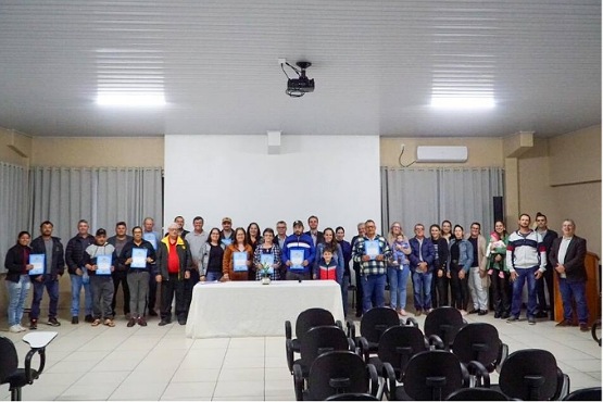 A imagem mostra um grupo grande de pessoas posando para uma foto em um ambiente interno, que parece ser uma sala de conferências ou auditório. Há cerca de 30 pessoas alinhadas em duas fileiras, uma atrás da outra. Na frente delas, há uma mesa coberta com uma toalha branca, e sobre a mesa há alguns objetos que não são claramente visíveis. Atrás do grupo, há uma tela de projeção branca e cortinas cinza nas laterais. O teto é branco com iluminação embutida e há um projetor pendurado no centro. Na frente da sala, há várias fileiras de cadeiras pretas vazias. As pessoas na foto estão vestidas de maneira casual e algumas seguram certificados ou documentos.