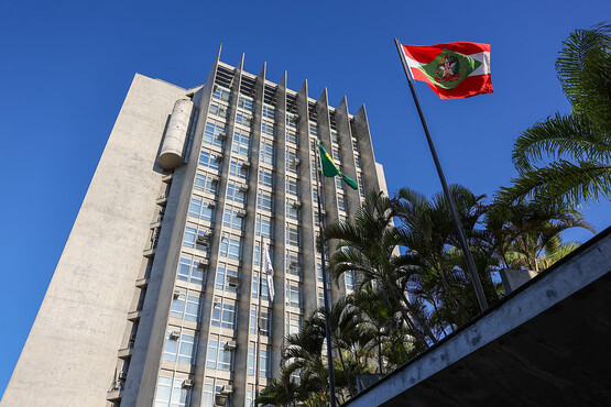 A imagem mostra um prédio alto de concreto com várias janelas, provavelmente um edifício governamental ou administrativo. Em frente ao prédio, há duas bandeiras hasteadas. A bandeira mais próxima é a do estado de Santa Catarina, que tem um fundo vermelho com um losango verde e um brasão no centro. A outra bandeira, um pouco mais distante, é a bandeira do Brasil. Ao redor das bandeiras, há várias palmeiras, e o céu está claro e azul, indicando um dia ensolarado.