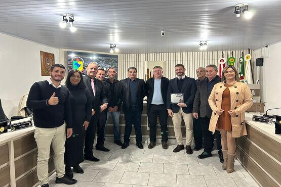 Onze pessoas, dentre elas homens e mulheres, estão posando para uma foto. Eles estão lado a lado dentro de uma sala que parece ser uma sala de reunião, há uma mesa em formato de U em volta deles. No fundo as paredes são beges e há bandeiras em hastes. O ambiente é bem iluminado.